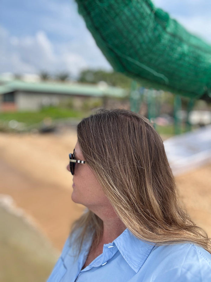 A candid side-profile photo of a woman standing near the shoreline, sunlight catching the soft waves in her hair. She’s wearing dark sunglasses and a light blue shirt, looking out toward the water with a calm, reflective expression. The blurred beach, net